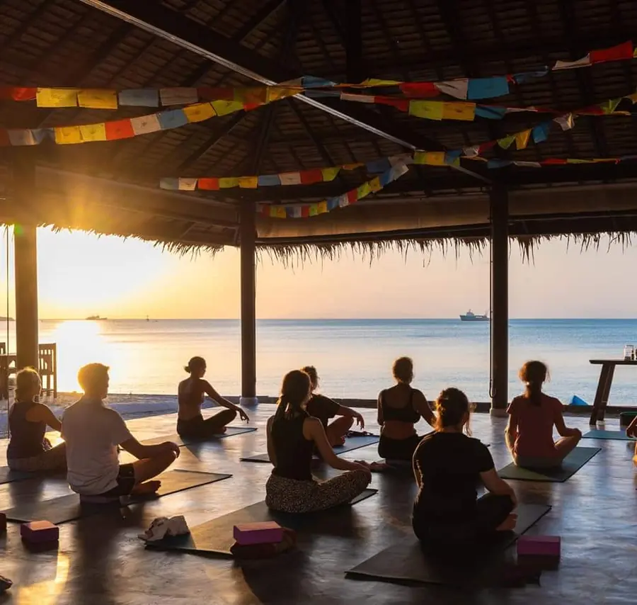 Group meditating in a beachfront yoga shala at sunset, prayer flags overhead.