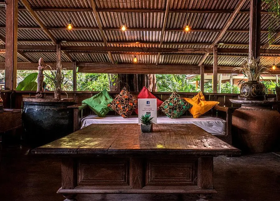Covered dining sala with wooden tables, Thai cushions and lanterns among tropical plants.