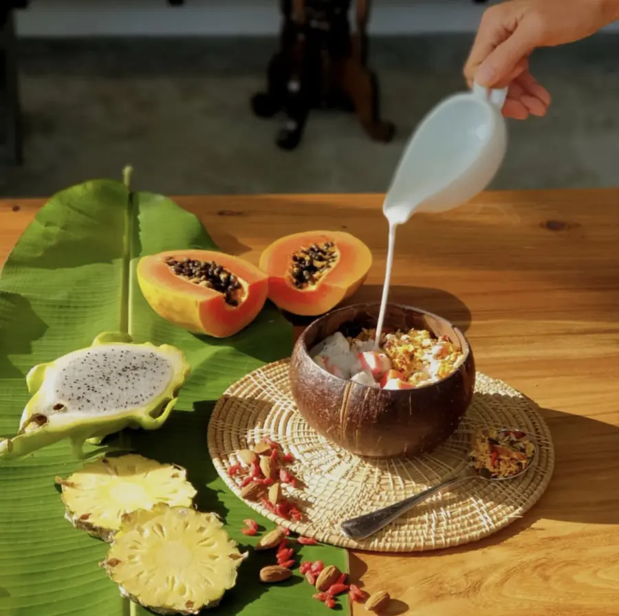 Coconut-shell bowl of granola and tropical fruit with coconut milk being poured over the top.
