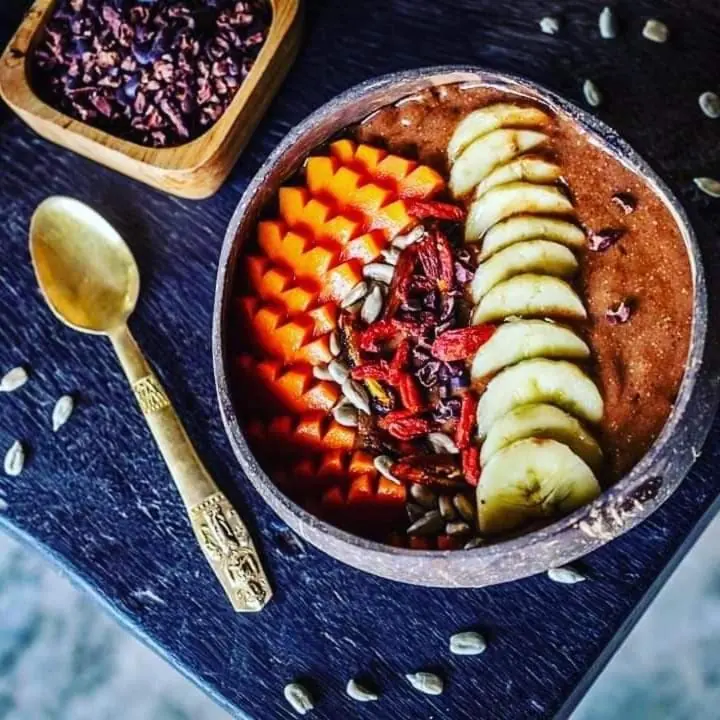 Smoothie bowl topped with sliced mango, banana and seeds, with cacao on the side.