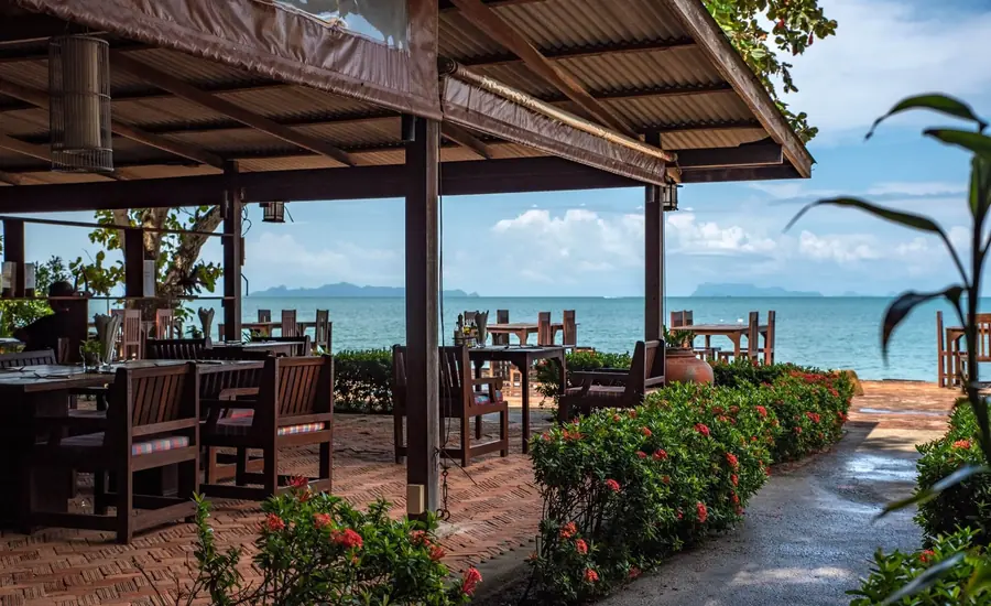 Open-air dining terrace with wooden tables facing the sea, flowering shrubs lining the path.
