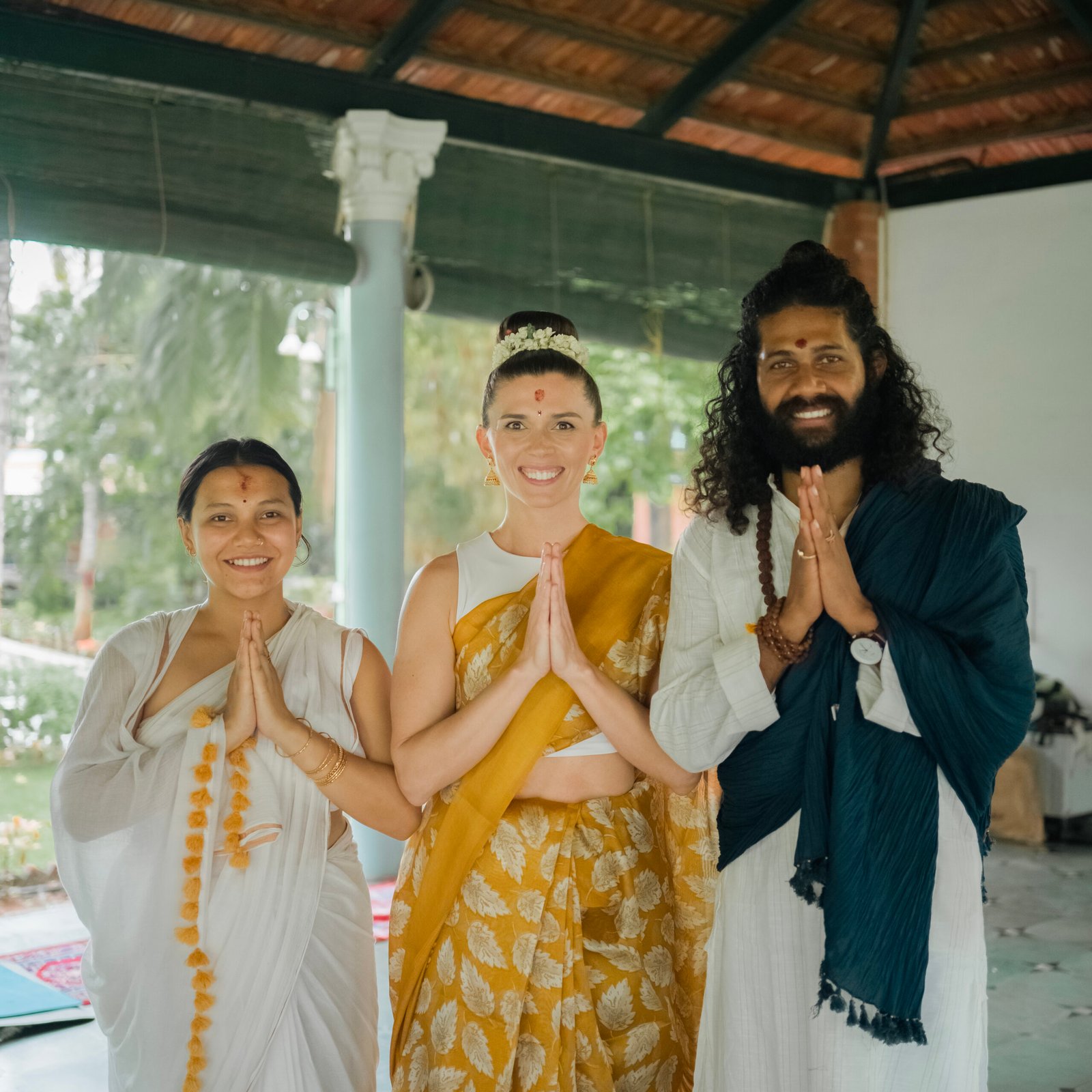 Three people in traditional Indian dress with marigold garlands greeting with namaste