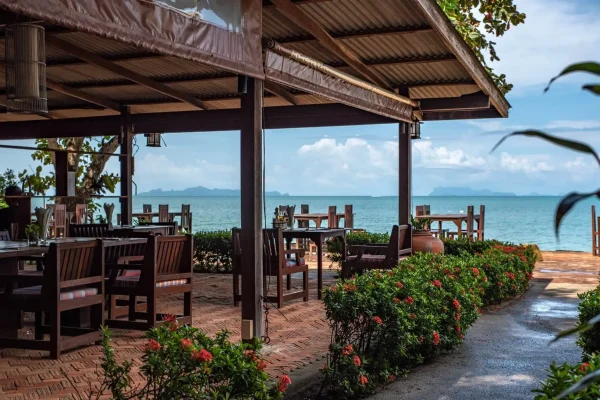 Open-air dining terrace with wooden tables and chairs facing the sea, flowering shrubs lining the path.